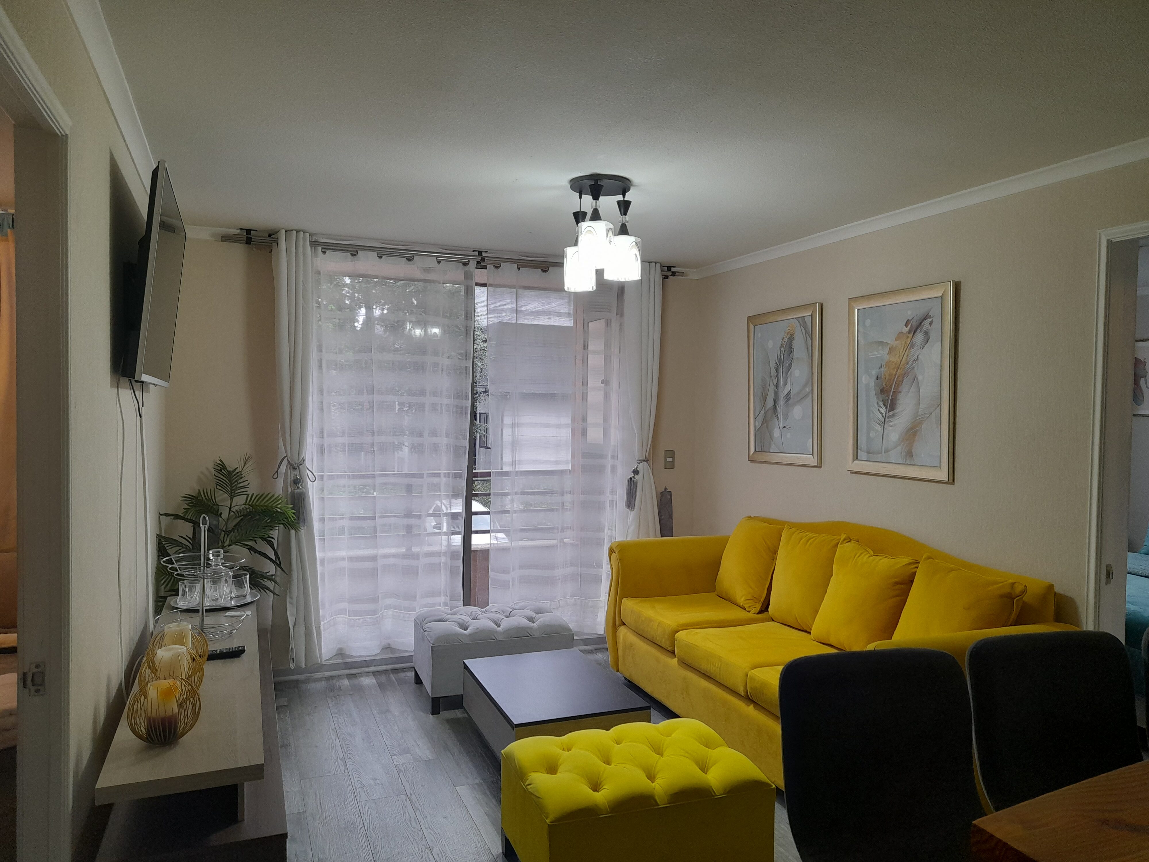 Sala de estar con sofá amarillo vibrante, mesa de centro moderna, cuadros decorativos en la pared y un gran ventanal con cortinas blancas que da hacia un balcón.